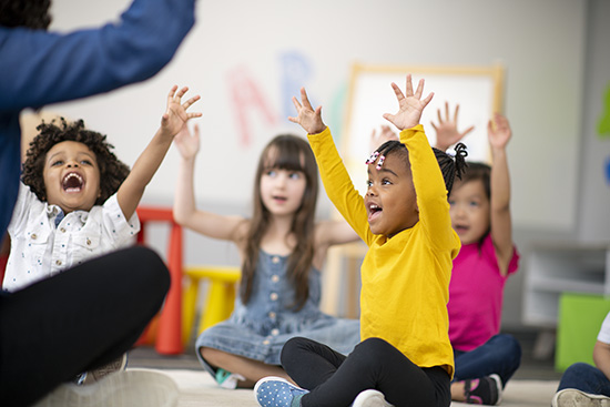 A multi-ethnic group of preschool students sitting on the floor in their classroom raising their hands.