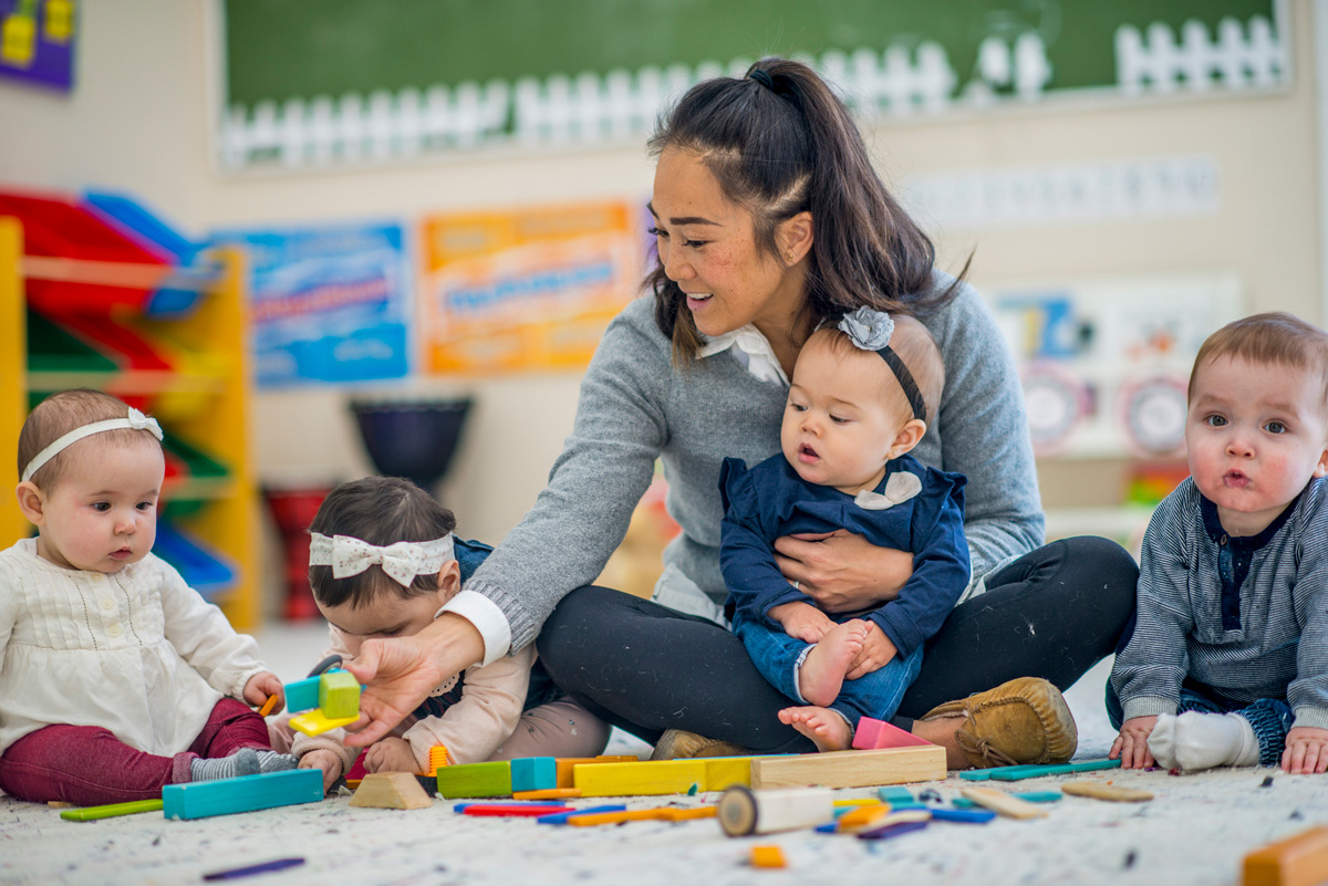 Caregiver sitting on a classroom floor with four infants, guiding them as they play with colorful blocks and toys.